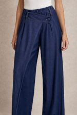 Viscose Denim Wrap Wide Leg Pants Χαρακτηριστικά Ύφασμα: Viscose με denim effect Υφή: Πολύ μαλακή και απαλή Αίσθηση: Ελαστική και άνετη Γραμμή: Wide leg Μέση: Ιδιαίτερη λοξή / asymmetric wrap μέση Λάστιχο στη μέση για μεγαλύτερη άνεση Λεπτομέρεια: Διακοσμητικό κουμπί στο μπροστινό μέρος Τσέπες: Πλαϊνές Εφαρμογή: Άνετη, με ρευστό πέσιμο Μέγεθος: One Size Καλύπτει έως Large Sensitive Skin Friendly Η απαλή viscose και η μαλακή αίσθηση του υφάσματος την καθιστούν κατάλληλη επιλογή για όσους προτιμούν μαλακά υφάσματα ή έχουν ευαίσθητο δέρμα. Σε αντίθεση με ένα πιο άκαμπτο denim, έχει πιο ήπια και ευχάριστη επαφή με το σώμα. Φροντίδα Πλύσιμο έως 30°C, σε χαμηλές στροφές. Αποφύγετε το έντονο στύψιμο. Εάν χρειαστεί σιδέρωμα, χρησιμοποιήστε χαμηλή θερμοκρασία. Προτείνεται φυσικό στέγνωμα και αποφυγή πολύωρης άμεσης έκθεσης στον ήλιο, ώστε να διατηρηθεί καλύτερα η απόχρωση και η υφή του υφάσματος.