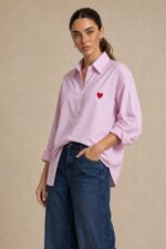 Cotton Oversized Heart Shirt  Χαρακτηριστικά Προϊόντος Ύφασμα: Μαλακό, σταθερό βαμβακερό ύφασμα Γραμμή: Oversized / Relaxed Fit Μέγεθος: One Size – καλύπτει έως Large Plus Ροζ βάση με λεπτές λευκές κάθετες ρίγες Κόκκινη κεντητή καρδιά στο μπροστινό μέρος Κλασικός γιακάς πουκαμίσου Κλείσιμο μπροστά με κουμπιά Μακριά μανίκια με μανσέτες Χαμηλή ραφή ώμων (dropped shoulders) Ασύμμετρο μήκος, πιο μακρύ πίσω Στρογγυλεμένο τελείωμα Πιέτα στην πλάτη για μεγαλύτερη άνεση Φαρδιά, αέρινη εφαρμογή Μέτρηση One Size Στήθος έως 125 cm Περιφέρεια έως 130 cm Οδηγίες Φροντίδας Πλύσιμο έως 30°C Χωρίς στύψιμο Πλύσιμο με παρόμοια χρώματα Προτείνεται πλύσιμο από την ανάποδη για προστασία του κεντήματος Φυσικό στέγνωμα σε σκιερό μέρος Όχι στεγνωτήριο Σιδέρωμα σε χαμηλή προς μέτρια θερμοκρασία, σύμφωνα με την ετικέτα σύνθεσης Αποφύγετε το απευθείας σιδέρωμα πάνω στην κεντητή καρδιά