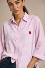 Cotton Oversized Heart Shirt  Χαρακτηριστικά Προϊόντος Ύφασμα: Μαλακό, σταθερό βαμβακερό ύφασμα Γραμμή: Oversized / Relaxed Fit Μέγεθος: One Size – καλύπτει έως Large Plus Ροζ βάση με λεπτές λευκές κάθετες ρίγες Κόκκινη κεντητή καρδιά στο μπροστινό μέρος Κλασικός γιακάς πουκαμίσου Κλείσιμο μπροστά με κουμπιά Μακριά μανίκια με μανσέτες Χαμηλή ραφή ώμων (dropped shoulders) Ασύμμετρο μήκος, πιο μακρύ πίσω Στρογγυλεμένο τελείωμα Πιέτα στην πλάτη για μεγαλύτερη άνεση Φαρδιά, αέρινη εφαρμογή Μέτρηση One Size Στήθος έως 125 cm Περιφέρεια έως 130 cm Οδηγίες Φροντίδας Πλύσιμο έως 30°C Χωρίς στύψιμο Πλύσιμο με παρόμοια χρώματα Προτείνεται πλύσιμο από την ανάποδη για προστασία του κεντήματος Φυσικό στέγνωμα σε σκιερό μέρος Όχι στεγνωτήριο Σιδέρωμα σε χαμηλή προς μέτρια θερμοκρασία, σύμφωνα με την ετικέτα σύνθεσης Αποφύγετε το απευθείας σιδέρωμα πάνω στην κεντητή καρδιά