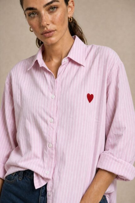 Cotton Oversized Heart Shirt  Χαρακτηριστικά Προϊόντος Ύφασμα: Μαλακό, σταθερό βαμβακερό ύφασμα Γραμμή: Oversized / Relaxed Fit Μέγεθος: One Size – καλύπτει έως Large Plus Ροζ βάση με λεπτές λευκές κάθετες ρίγες Κόκκινη κεντητή καρδιά στο μπροστινό μέρος Κλασικός γιακάς πουκαμίσου Κλείσιμο μπροστά με κουμπιά Μακριά μανίκια με μανσέτες Χαμηλή ραφή ώμων (dropped shoulders) Ασύμμετρο μήκος, πιο μακρύ πίσω Στρογγυλεμένο τελείωμα Πιέτα στην πλάτη για μεγαλύτερη άνεση Φαρδιά, αέρινη εφαρμογή Μέτρηση One Size Στήθος έως 125 cm Περιφέρεια έως 130 cm Οδηγίες Φροντίδας Πλύσιμο έως 30°C Χωρίς στύψιμο Πλύσιμο με παρόμοια χρώματα Προτείνεται πλύσιμο από την ανάποδη για προστασία του κεντήματος Φυσικό στέγνωμα σε σκιερό μέρος Όχι στεγνωτήριο Σιδέρωμα σε χαμηλή προς μέτρια θερμοκρασία, σύμφωνα με την ετικέτα σύνθεσης Αποφύγετε το απευθείας σιδέρωμα πάνω στην κεντητή καρδιά