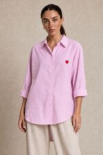 Cotton Oversized Heart Shirt  Χαρακτηριστικά Προϊόντος Ύφασμα: Μαλακό, σταθερό βαμβακερό ύφασμα Γραμμή: Oversized / Relaxed Fit Μέγεθος: One Size – καλύπτει έως Large Plus Ροζ βάση με λεπτές λευκές κάθετες ρίγες Κόκκινη κεντητή καρδιά στο μπροστινό μέρος Κλασικός γιακάς πουκαμίσου Κλείσιμο μπροστά με κουμπιά Μακριά μανίκια με μανσέτες Χαμηλή ραφή ώμων (dropped shoulders) Ασύμμετρο μήκος, πιο μακρύ πίσω Στρογγυλεμένο τελείωμα Πιέτα στην πλάτη για μεγαλύτερη άνεση Φαρδιά, αέρινη εφαρμογή Μέτρηση One Size Στήθος έως 125 cm Περιφέρεια έως 130 cm Οδηγίες Φροντίδας Πλύσιμο έως 30°C Χωρίς στύψιμο Πλύσιμο με παρόμοια χρώματα Προτείνεται πλύσιμο από την ανάποδη για προστασία του κεντήματος Φυσικό στέγνωμα σε σκιερό μέρος Όχι στεγνωτήριο Σιδέρωμα σε χαμηλή προς μέτρια θερμοκρασία, σύμφωνα με την ετικέτα σύνθεσης Αποφύγετε το απευθείας σιδέρωμα πάνω στην κεντητή καρδιά