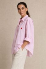 Cotton Oversized Heart Shirt  Χαρακτηριστικά Προϊόντος Ύφασμα: Μαλακό, σταθερό βαμβακερό ύφασμα Γραμμή: Oversized / Relaxed Fit Μέγεθος: One Size – καλύπτει έως Large Plus Ροζ βάση με λεπτές λευκές κάθετες ρίγες Κόκκινη κεντητή καρδιά στο μπροστινό μέρος Κλασικός γιακάς πουκαμίσου Κλείσιμο μπροστά με κουμπιά Μακριά μανίκια με μανσέτες Χαμηλή ραφή ώμων (dropped shoulders) Ασύμμετρο μήκος, πιο μακρύ πίσω Στρογγυλεμένο τελείωμα Πιέτα στην πλάτη για μεγαλύτερη άνεση Φαρδιά, αέρινη εφαρμογή Μέτρηση One Size Στήθος έως 125 cm Περιφέρεια έως 130 cm Οδηγίες Φροντίδας Πλύσιμο έως 30°C Χωρίς στύψιμο Πλύσιμο με παρόμοια χρώματα Προτείνεται πλύσιμο από την ανάποδη για προστασία του κεντήματος Φυσικό στέγνωμα σε σκιερό μέρος Όχι στεγνωτήριο Σιδέρωμα σε χαμηλή προς μέτρια θερμοκρασία, σύμφωνα με την ετικέτα σύνθεσης Αποφύγετε το απευθείας σιδέρωμα πάνω στην κεντητή καρδιά