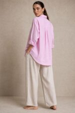 Cotton Oversized Heart Shirt  Χαρακτηριστικά Προϊόντος Ύφασμα: Μαλακό, σταθερό βαμβακερό ύφασμα Γραμμή: Oversized / Relaxed Fit Μέγεθος: One Size – καλύπτει έως Large Plus Ροζ βάση με λεπτές λευκές κάθετες ρίγες Κόκκινη κεντητή καρδιά στο μπροστινό μέρος Κλασικός γιακάς πουκαμίσου Κλείσιμο μπροστά με κουμπιά Μακριά μανίκια με μανσέτες Χαμηλή ραφή ώμων (dropped shoulders) Ασύμμετρο μήκος, πιο μακρύ πίσω Στρογγυλεμένο τελείωμα Πιέτα στην πλάτη για μεγαλύτερη άνεση Φαρδιά, αέρινη εφαρμογή Μέτρηση One Size Στήθος έως 125 cm Περιφέρεια έως 130 cm Οδηγίες Φροντίδας Πλύσιμο έως 30°C Χωρίς στύψιμο Πλύσιμο με παρόμοια χρώματα Προτείνεται πλύσιμο από την ανάποδη για προστασία του κεντήματος Φυσικό στέγνωμα σε σκιερό μέρος Όχι στεγνωτήριο Σιδέρωμα σε χαμηλή προς μέτρια θερμοκρασία, σύμφωνα με την ετικέτα σύνθεσης Αποφύγετε το απευθείας σιδέρωμα πάνω στην κεντητή καρδιά