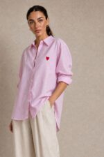 Cotton Oversized Heart Shirt  Χαρακτηριστικά Προϊόντος Ύφασμα: Μαλακό, σταθερό βαμβακερό ύφασμα Γραμμή: Oversized / Relaxed Fit Μέγεθος: One Size – καλύπτει έως Large Plus Ροζ βάση με λεπτές λευκές κάθετες ρίγες Κόκκινη κεντητή καρδιά στο μπροστινό μέρος Κλασικός γιακάς πουκαμίσου Κλείσιμο μπροστά με κουμπιά Μακριά μανίκια με μανσέτες Χαμηλή ραφή ώμων (dropped shoulders) Ασύμμετρο μήκος, πιο μακρύ πίσω Στρογγυλεμένο τελείωμα Πιέτα στην πλάτη για μεγαλύτερη άνεση Φαρδιά, αέρινη εφαρμογή Μέτρηση One Size Στήθος έως 125 cm Περιφέρεια έως 130 cm Οδηγίες Φροντίδας Πλύσιμο έως 30°C Χωρίς στύψιμο Πλύσιμο με παρόμοια χρώματα Προτείνεται πλύσιμο από την ανάποδη για προστασία του κεντήματος Φυσικό στέγνωμα σε σκιερό μέρος Όχι στεγνωτήριο Σιδέρωμα σε χαμηλή προς μέτρια θερμοκρασία, σύμφωνα με την ετικέτα σύνθεσης Αποφύγετε το απευθείας σιδέρωμα πάνω στην κεντητή καρδιά