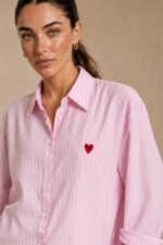 Cotton Oversized Heart Shirt  Χαρακτηριστικά Προϊόντος Ύφασμα: Μαλακό, σταθερό βαμβακερό ύφασμα Γραμμή: Oversized / Relaxed Fit Μέγεθος: One Size – καλύπτει έως Large Plus Ροζ βάση με λεπτές λευκές κάθετες ρίγες Κόκκινη κεντητή καρδιά στο μπροστινό μέρος Κλασικός γιακάς πουκαμίσου Κλείσιμο μπροστά με κουμπιά Μακριά μανίκια με μανσέτες Χαμηλή ραφή ώμων (dropped shoulders) Ασύμμετρο μήκος, πιο μακρύ πίσω Στρογγυλεμένο τελείωμα Πιέτα στην πλάτη για μεγαλύτερη άνεση Φαρδιά, αέρινη εφαρμογή Μέτρηση One Size Στήθος έως 125 cm Περιφέρεια έως 130 cm Οδηγίες Φροντίδας Πλύσιμο έως 30°C Χωρίς στύψιμο Πλύσιμο με παρόμοια χρώματα Προτείνεται πλύσιμο από την ανάποδη για προστασία του κεντήματος Φυσικό στέγνωμα σε σκιερό μέρος Όχι στεγνωτήριο Σιδέρωμα σε χαμηλή προς μέτρια θερμοκρασία, σύμφωνα με την ετικέτα σύνθεσης Αποφύγετε το απευθείας σιδέρωμα πάνω στην κεντητή καρδιά