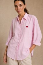 Cotton Oversized Heart Shirt  Χαρακτηριστικά Προϊόντος Ύφασμα: Μαλακό, σταθερό βαμβακερό ύφασμα Γραμμή: Oversized / Relaxed Fit Μέγεθος: One Size – καλύπτει έως Large Plus Ροζ βάση με λεπτές λευκές κάθετες ρίγες Κόκκινη κεντητή καρδιά στο μπροστινό μέρος Κλασικός γιακάς πουκαμίσου Κλείσιμο μπροστά με κουμπιά Μακριά μανίκια με μανσέτες Χαμηλή ραφή ώμων (dropped shoulders) Ασύμμετρο μήκος, πιο μακρύ πίσω Στρογγυλεμένο τελείωμα Πιέτα στην πλάτη για μεγαλύτερη άνεση Φαρδιά, αέρινη εφαρμογή Μέτρηση One Size Στήθος έως 125 cm Περιφέρεια έως 130 cm Οδηγίες Φροντίδας Πλύσιμο έως 30°C Χωρίς στύψιμο Πλύσιμο με παρόμοια χρώματα Προτείνεται πλύσιμο από την ανάποδη για προστασία του κεντήματος Φυσικό στέγνωμα σε σκιερό μέρος Όχι στεγνωτήριο Σιδέρωμα σε χαμηλή προς μέτρια θερμοκρασία, σύμφωνα με την ετικέτα σύνθεσης Αποφύγετε το απευθείας σιδέρωμα πάνω στην κεντητή καρδιά